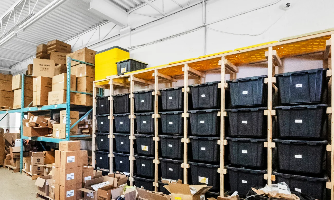 Labelled black storage bins in wooden racks beside teal pallet racking stacked with cartons